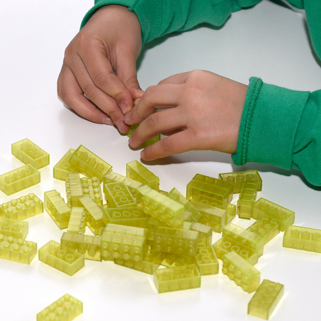 Hands joining yellow 2x4 Building Bricks, many bricks scattered on a white surface