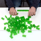 Hands laying a row of green 2x4 building bricks, more bricks scattered on a white table
