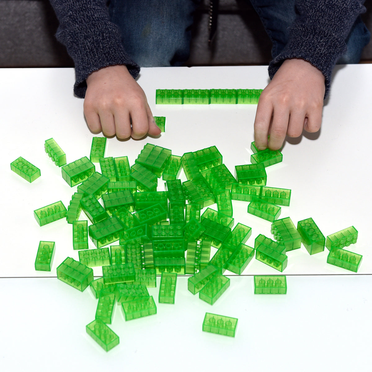 Hands laying a row of green 2x4 building bricks, more bricks scattered on a white table