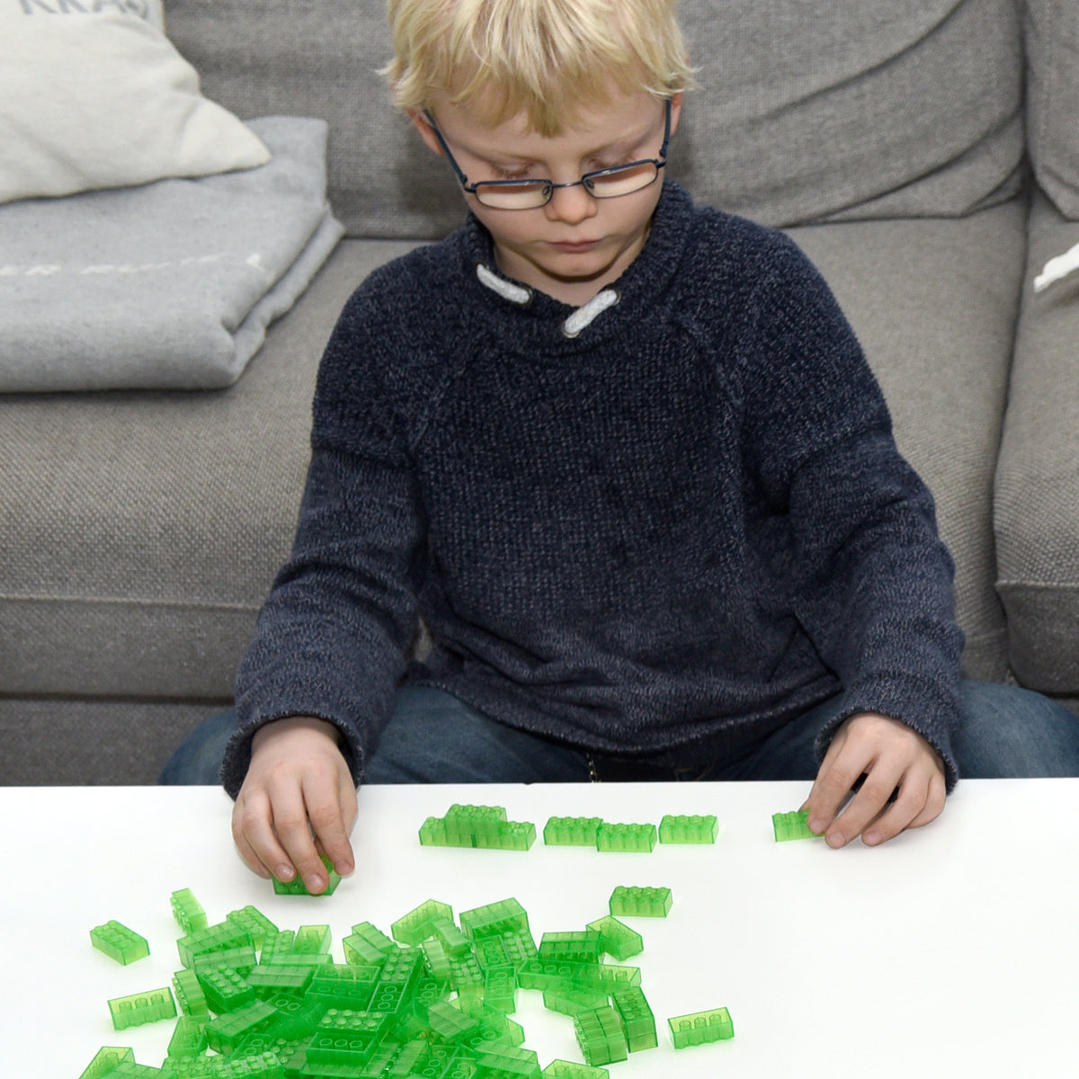Child wearing glasses sitting in front of a sofa and lining up green 2x4 building bricks on a white table
