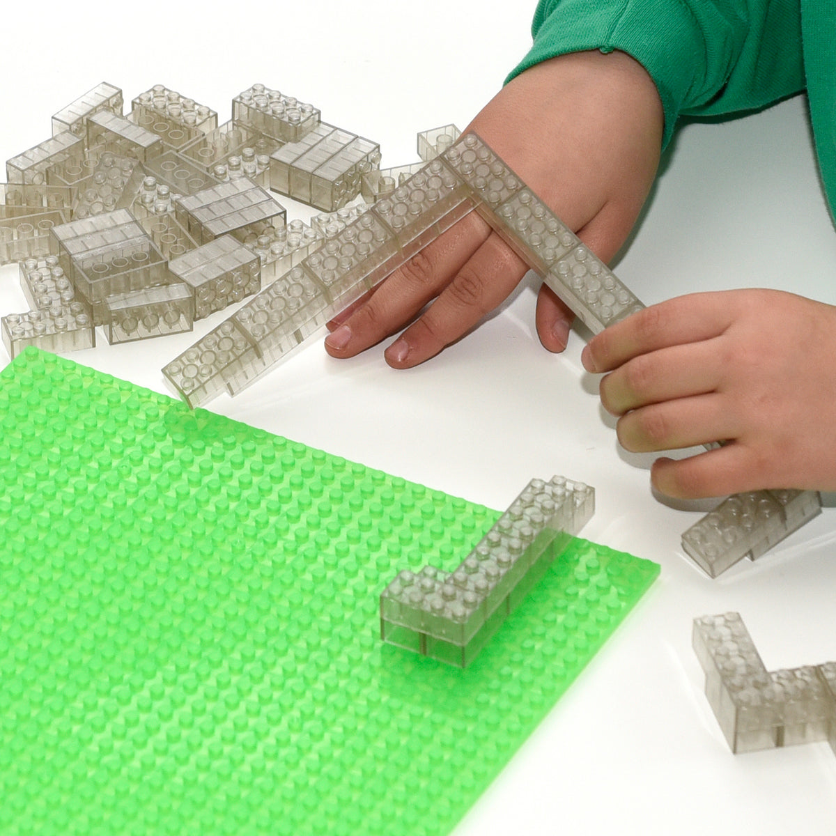 Transparent 2x4 building bricks on a green baseplate, children's hands building